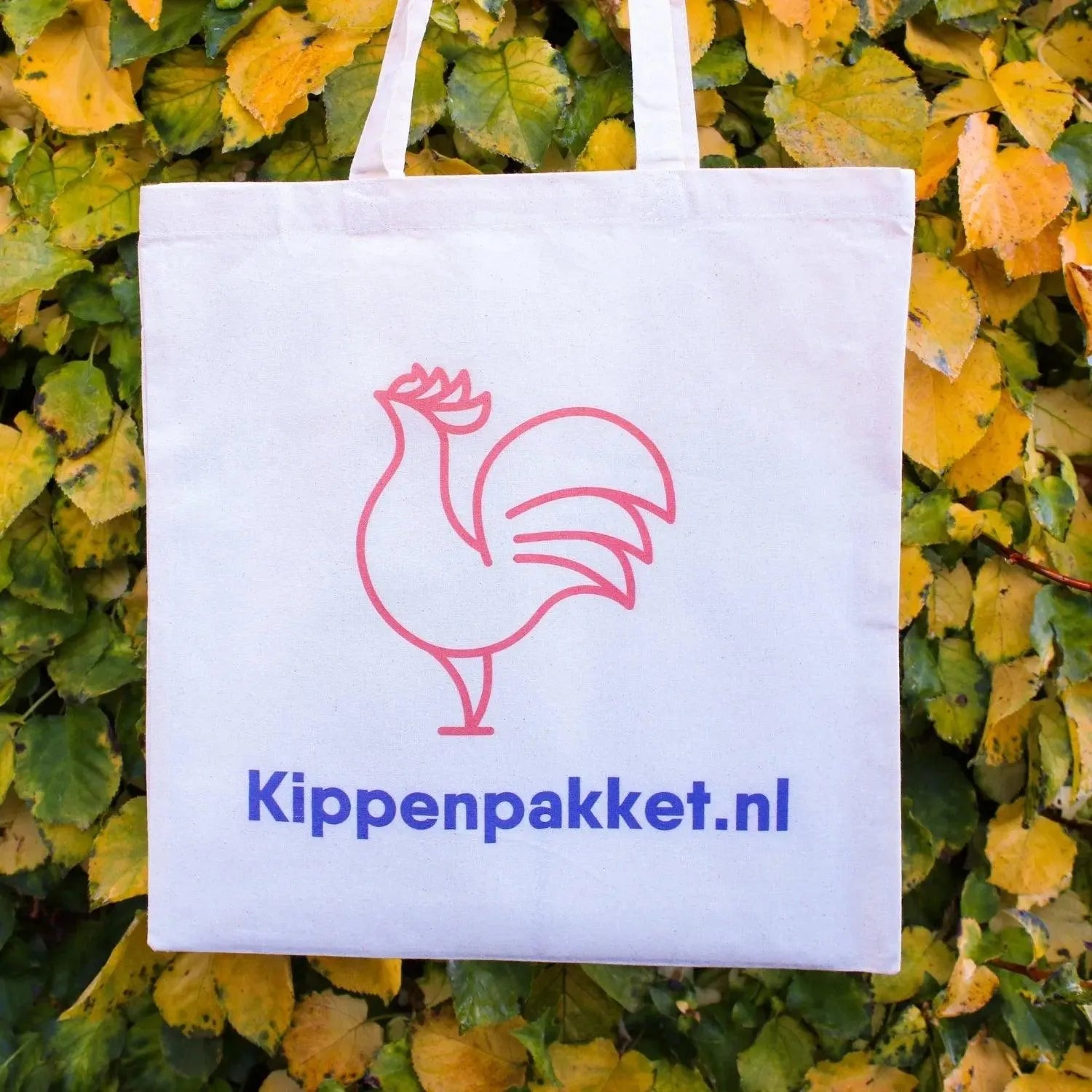 Een witte Kippenpakket Tas van Kippenpakket.nl heeft een rode haanlijntekening met "Kippenpakket.nl" in het blauw. Ideaal voor boodschappen, de tas is afgezet tegen een levendige groene en gele bladachtergrond.