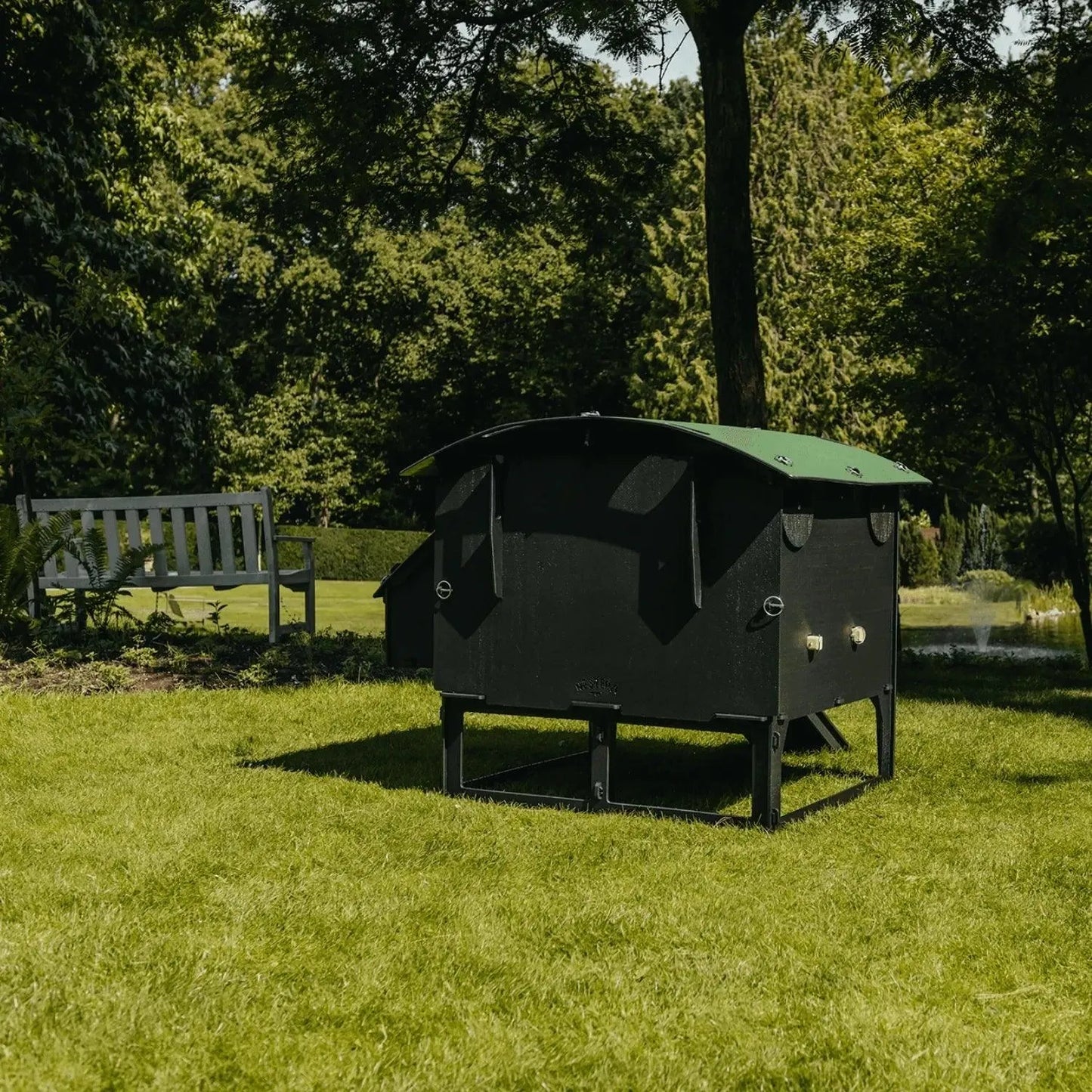 Een grote zwart-groene houten schuilplaats, die doet denken aan de Nestera kunststof kippenhok op poten van Kippenpakket.nl, staat in het grasveld van een park. Op de achtergrond staat een bankje tussen bomen en struiken met een verre vijver in het zicht.