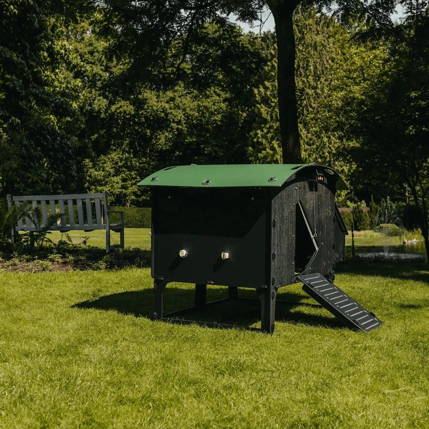 Een Nestera kunststof kippenhok op poten large van Kippenpakket.nl, met een groen dak, staat op een grasveld. Een kleine helling leidt naar de ingang, ontworpen om bloedluizen te weren. Op de achtergrond zorgen een houten bank en bomen voor schaduw.