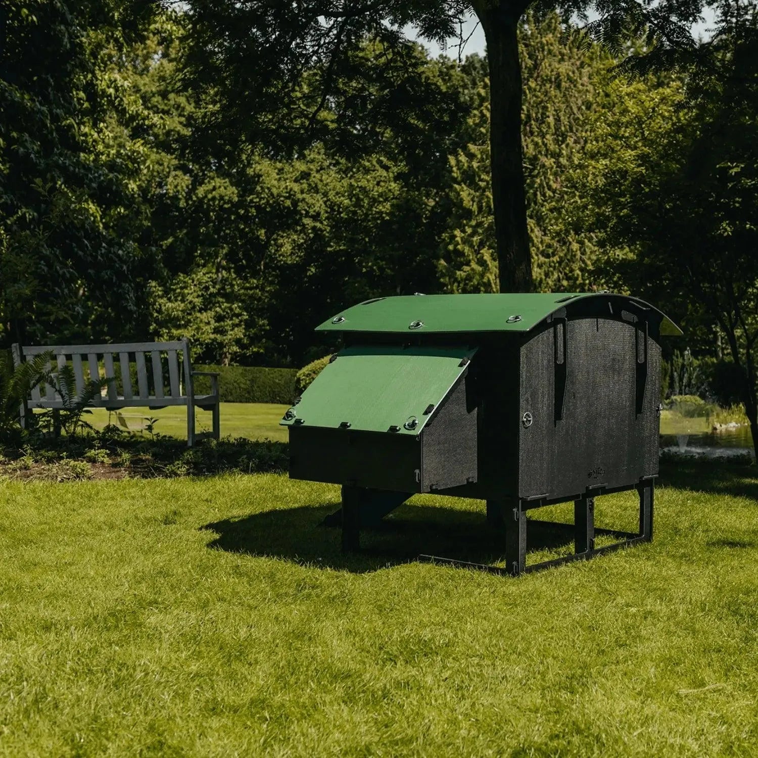 Een Nestera kunststof kippenhok op poten large van Kippenpakket.nl, geschikt voor 4 tot 10 kippen, staat op een grasveld met bomen. Een wit bankje bij een vijver versterkt de zonnige setting. Dit hok, in het zwart met een groen dak, is gemaakt van duurzaam gerecycled kunststof om bloedluizen efficiënt te voorkomen.