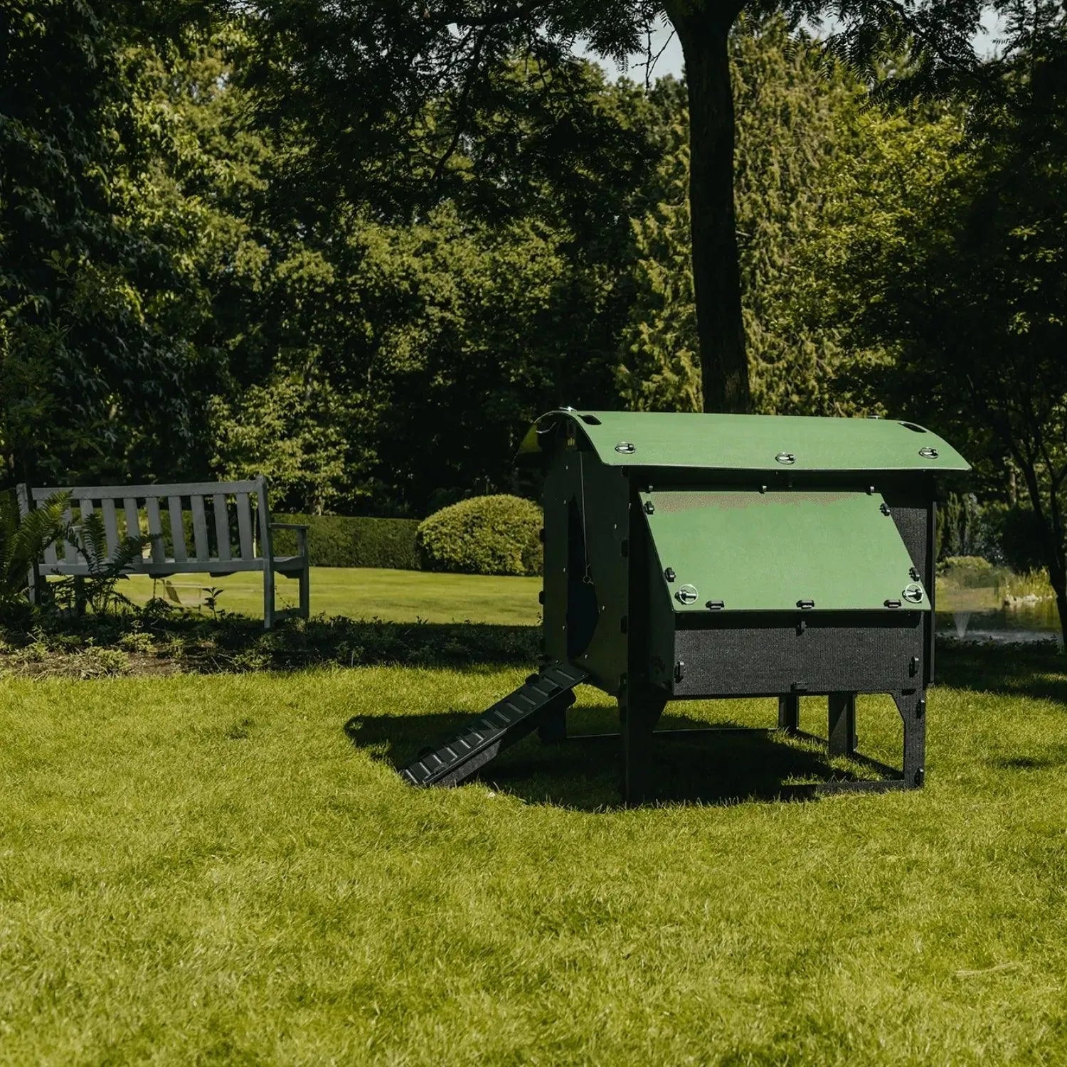 Een Nestera kunststof kippenhok op poten large van Kippenpakket.nl, gemaakt om bloedluizen te voorkomen en vervaardigd uit duurzaam gerecycled kunststof, staat op een grasveld in een zonnige tuin. Tussen bomen en struiken onder een helderblauwe lucht is een houten bank zichtbaar.