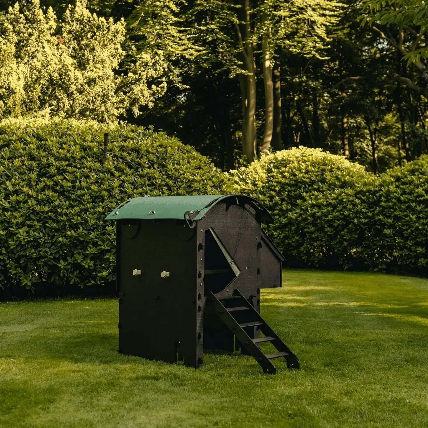 Het Nestera kunststof kippenhok op poten medium van Kippenpakket.nl, geschikt voor 3 tot 9 kippen, staat op een verzorgd gazon. Met donkergroene en zwarte panelen, een gebogen dak en ladder is dit bloedluisbestendig hok omgeven door struiken, gras en bomen bij daglicht - functioneel en charmant.